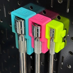 May include: Three metal socket extensions held by bright teal, hot pink, and neon yellow plastic organizers mounted on a black pegboard. Each organizer features a hexagonal logo on the top surface and holds a silver-colored steel tool extension.