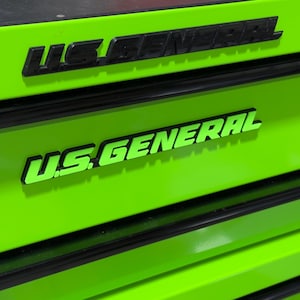 May include: Close-up of a bright green tool chest with black accents. The words "U.S. GENERAL" are prominently displayed in black and green lettering. The tool chest has multiple drawers and a black top.