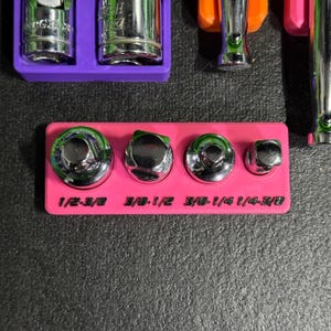 May include: A bright pink rectangular organiser tray holding four chrome metal socket adapters. The tray features black embossed text labels for each adapter size: 1/2-3/8, 3/8-1/2, 3/8-1/4, and 1/4-3/8. The adapters are arranged in a row.
