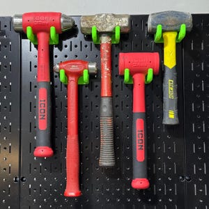 May include: Five different hammers hanging on a black pegboard wall. The hammers are all red and black with green and black holders. The hammers are labeled "ICON" and "ESTWING".