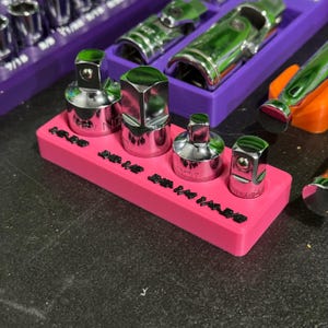 May include: A bright pink rectangular plastic organiser tray holding four chrome-plated metal socket adapters of varying sizes. The tray features embossed black text labels reading 1/4, 3/8, 3/8, and 1/4 in a row along the front edge of the base.