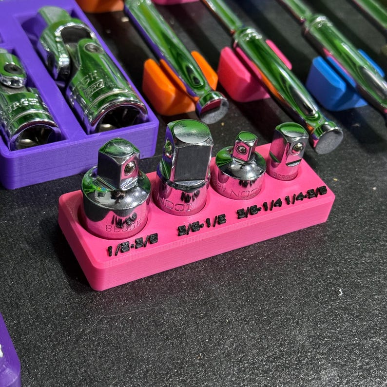 May include: A bright pink rectangular plastic organiser tray holding four chrome steel socket adapters. The tray features black embossed text labels reading 1/2-3/8, 3/8-1/2, 3/8-1/4, and 1/4-3/8. The adapters have square drive ends and polished metallic surfaces.