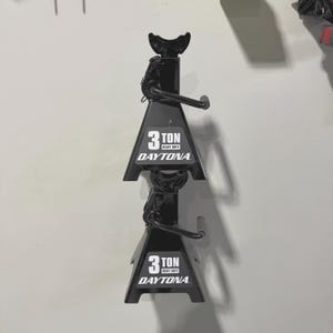 May include: Two black Daytona 3-ton heavy-duty jack stands. Each stand has a triangular base and a curved top for supporting vehicles. The Daytona logo is printed in white on the front of each stand.