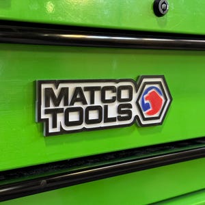 May include: Close-up of a green tool chest with a black and white "MATCO TOOLS" logo. The logo includes a red, white, and blue emblem. The tool chest has black trim and a lock.