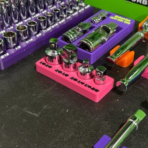 May include: A pink rectangular plastic organiser tray holding four chrome-plated metal socket adapters. The tray features black embossed text labels for each tool size. Additional purple organisers with various chrome sockets are visible nearby.