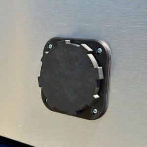 May include: A black circular cap with a notched edge, mounted on a square base plate. The assembly is secured to a brushed metallic surface with four silver-colored screws positioned at each corner of the square mounting plate.