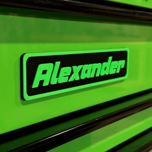 May include: A close-up of a bright green surface with a black rectangular label. The label features the name "Alexander" in bold, neon green letters. The background is a matching bright green.