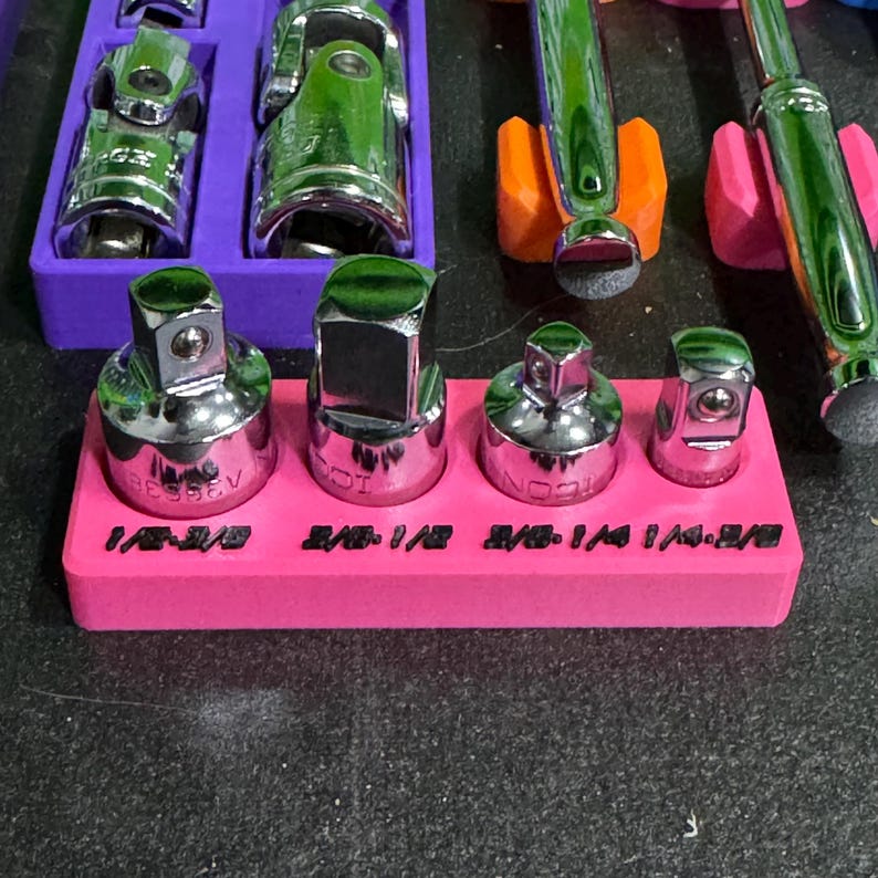 May include: A bright pink rectangular organiser tray holding four chrome metal socket adapters. The tray features black printed text labels for each adapter size, including 1/2-3/8, 3/8-1/2, 3/8-1/4, and 1/4-3/8, arranged in a single row.