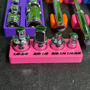 May include: A set of four chrome-plated steel socket adapters arranged in a bright pink plastic holder. The holder features black embossed text labels reading 1/2-3/8, 3/8-1/2, 3/8-1/4, and 1/4-3/8, indicating the specific drive size conversions for each tool.