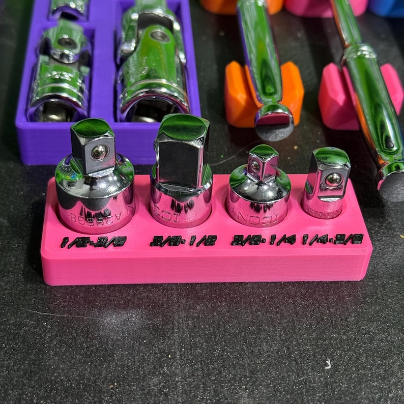 May include: A set of four chrome-plated steel socket adapters arranged in a bright pink plastic holder. The holder features black embossed text labels reading 1/2-3/8, 3/8-1/2, 3/8-1/4, and 1/4-3/8, indicating the specific drive size conversions for each tool.
