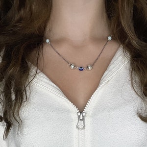 May include: A silver chain necklace with a delicate design. The necklace features small, round, silver beads, and a blue and white evil eye charm. The necklace is worn against a white background.