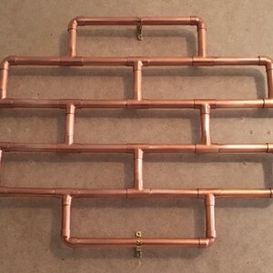 May include: A copper pipe wall shelf with a geometric design. The shelf is made of copper pipes and fittings and has a rustic industrial look.