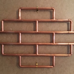 May include: A copper pipe wall shelf with a geometric design. The shelf has four rows of pipes and is mounted to the wall with four brass hooks.