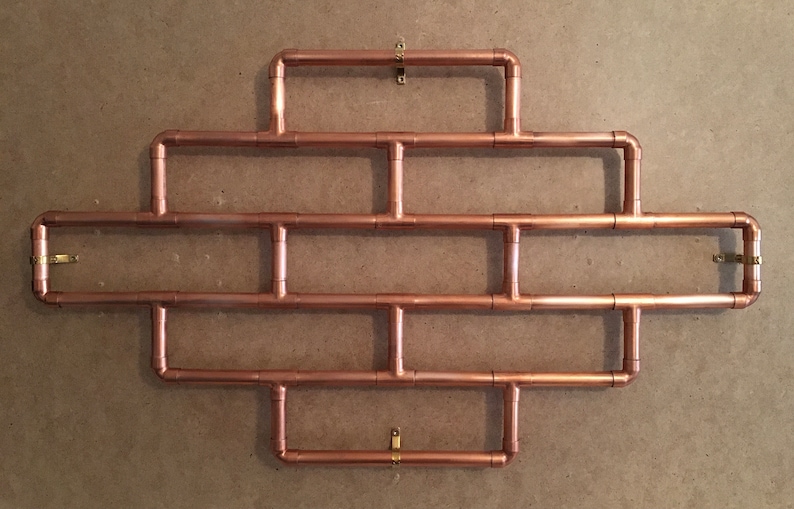 May include: A copper pipe wall shelf with a geometric design. The shelf has four rows of pipes and is mounted to the wall with four brass hooks.