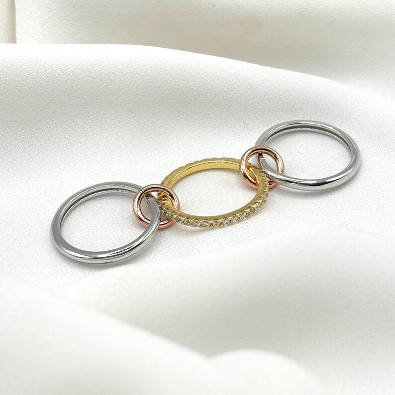 May include: Three interlocking rings, one gold with a row of small clear stones, one silver, and one rose gold.