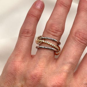 May include: A silver, gold, and rose gold ring with a row of clear stones. The ring is designed with three interlocking bands.