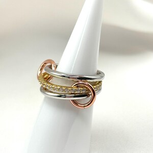 May include: A multi-layered ring with a silver, gold, and rose gold band. The ring features a row of small clear stones set in the gold band.