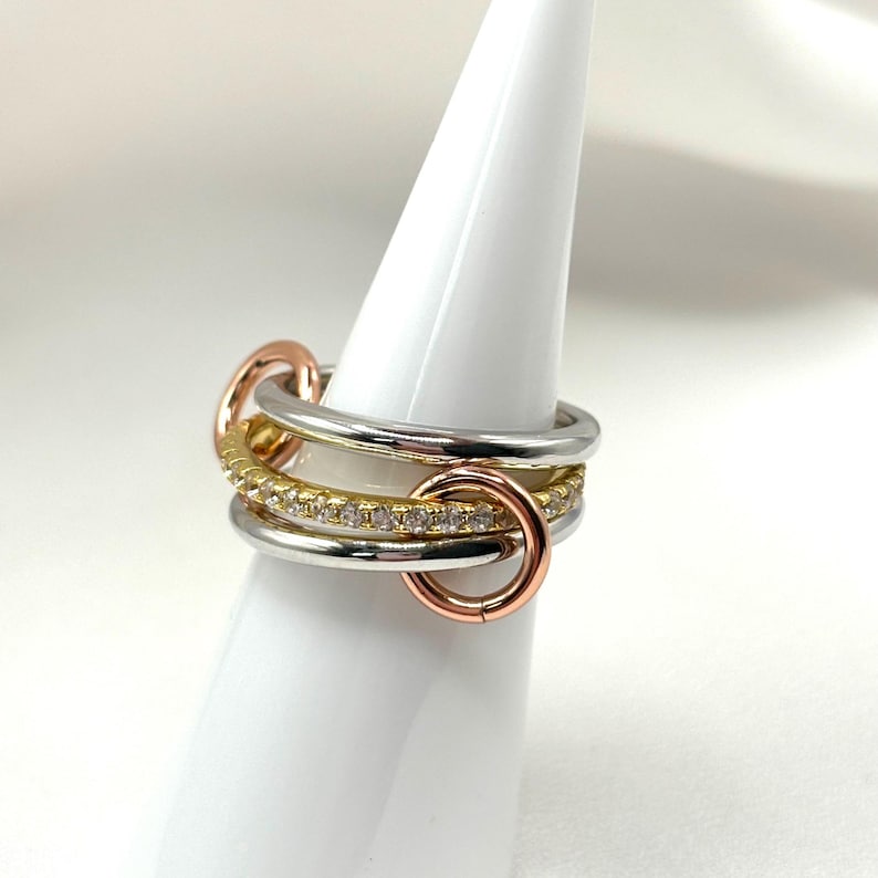 May include: A multi-layered ring with a silver, gold, and rose gold band. The ring features a row of small clear stones set in the gold band.