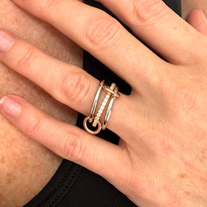 May include: A ring with three bands, two silver and one rose gold, with a row of small clear stones on the rose gold band. The bands are intertwined and the rose gold band has a small circle at the end.