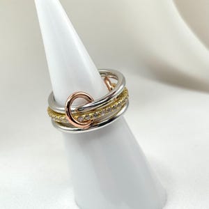 May include: A multi-layered ring with a gold band, a silver band, and a rose gold band with a circle design. The ring is set with small diamonds.