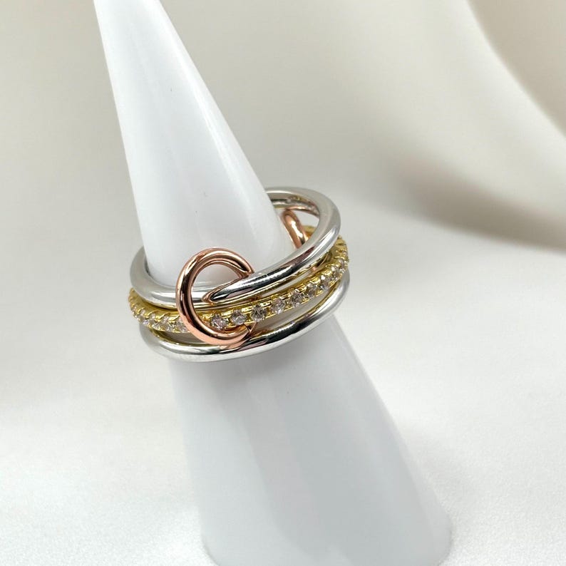 May include: A multi-layered ring with a gold band, a silver band, and a rose gold band with a circle design. The ring is set with small diamonds.