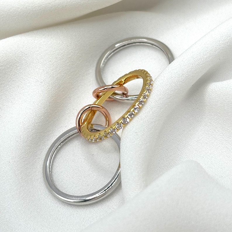May include: Three interlocking rings, one with a row of clear stones, on a white fabric background. The rings are gold, rose gold, and silver.