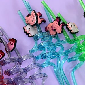 Colorful reusable drinking straws with axolotl toppers. The straws are in shades of pink, orange, green, purple, and yellow. The axolotl toppers are in matching colors. The background is a solid pink.