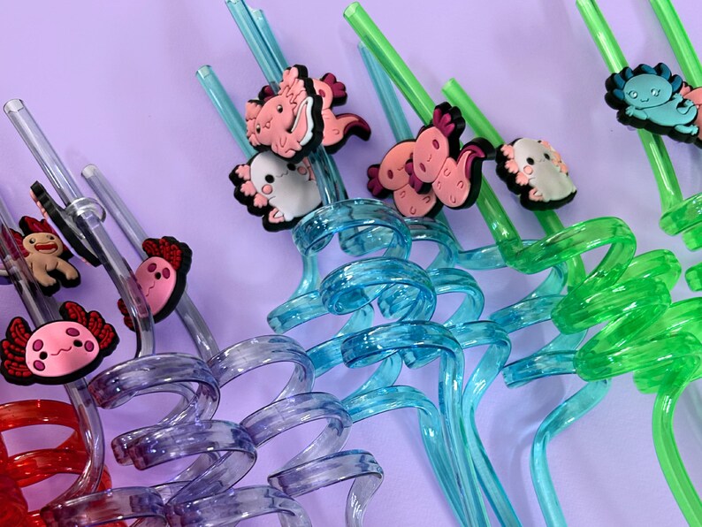 Colorful reusable drinking straws with axolotl toppers. The straws are in shades of pink, orange, green, purple, and yellow. The axolotl toppers are in matching colors. The background is a solid pink.