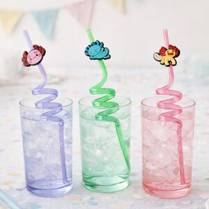 May include: Three clear glasses filled with ice and water, each with a colorful, decorative straw. The straws are purple, green, and pink, topped with cartoon animal charms. The glasses are on a white surface with pastel decorations.