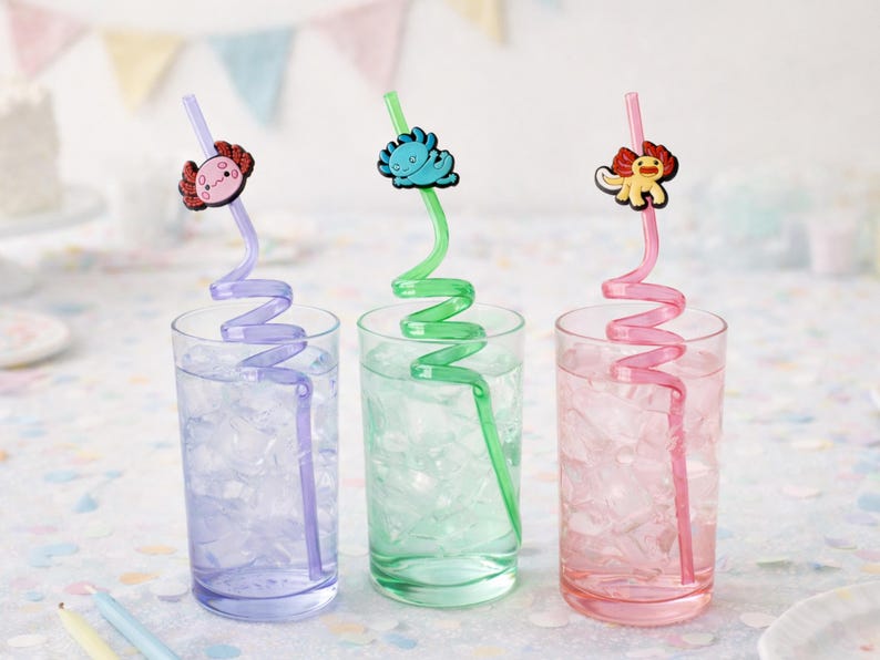 May include: Three clear glasses filled with ice and water, each with a colorful, decorative straw. The straws are purple, green, and pink, topped with cartoon animal charms. The glasses are on a white surface with pastel decorations.