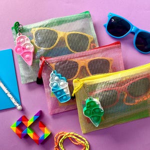 May include: An assortment of summer accessories, including sunglasses, mesh zipper pouches, and colorful pop-it keychains. A blue notebook, pen, and a rainbow puzzle are also present. The items are arranged on a purple surface.