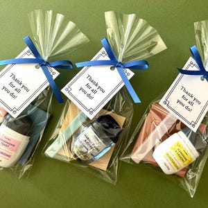 May include: Three clear cellophane gift bags tied with blue ribbon, each containing a small bottle of hand sanitizer and other items. Each bag has a tag that reads "Thank you for all you do!" The hand sanitizers are labeled "Champagne Toast", "Marine Faves & Citrus", and "Sunshine & Lemons".