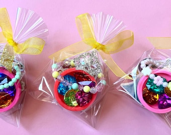 May include: Three clear plastic favor bags each containing a small pink plastic bowl filled with assorted colorful faceted plastic gems and a beaded bracelet with a silver-toned crown charm, all tied at the top with a yellow organza ribbon.