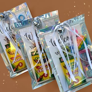 May include: Three clear, iridescent zipper bags filled with party favors. Each bag has a small disco ball, white ribbon, and a card that says "Welcome". Contents include crayons, toys, and a blue hair tie.