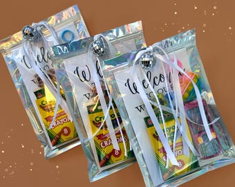 May include: Three clear, iridescent zipper bags filled with party favors. Each bag has a small disco ball, white ribbon, and a card that says "Welcome". Contents include crayons, toys, and a blue hair tie.