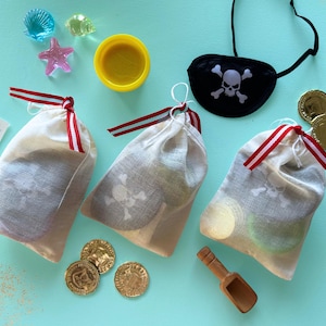 May include: Three off-white drawstring muslin bags with red and white striped ribbons, each containing a skull and crossbones print. Accessories include a black pirate eye patch, gold plastic coins, a small wooden scoop, and colorful sea-themed charms.