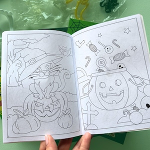 Puede incluir: Un libro para colorear abierto con dibujos de l&iacute;neas en blanco y negro. La p&aacute;gina izquierda muestra un gato con sombrero de bruja sentado sobre una calabaza. La p&aacute;gina derecha muestra un cubo de calabaza lleno de dulces, piruletas y estrellas.