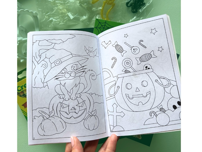 Puede incluir: Un libro para colorear abierto con dibujos de l&iacute;neas en blanco y negro. La p&aacute;gina izquierda muestra un gato con sombrero de bruja sentado sobre una calabaza. La p&aacute;gina derecha muestra un cubo de calabaza lleno de dulces, piruletas y estrellas.