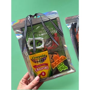 Puede incluir: Una bolsa de pl&aacute;stico iridiscente transparente atada con una cinta de rayas blancas y negras que contiene un libro para colorear de Happy Halloween, una caja peque&ntilde;a de ocho crayones Crayola, un marco de pl&aacute;stico blanco, un esqueleto de pl&aacute;stico rojo y un juguete fidget naranja con forma de murci&eacute;lago.