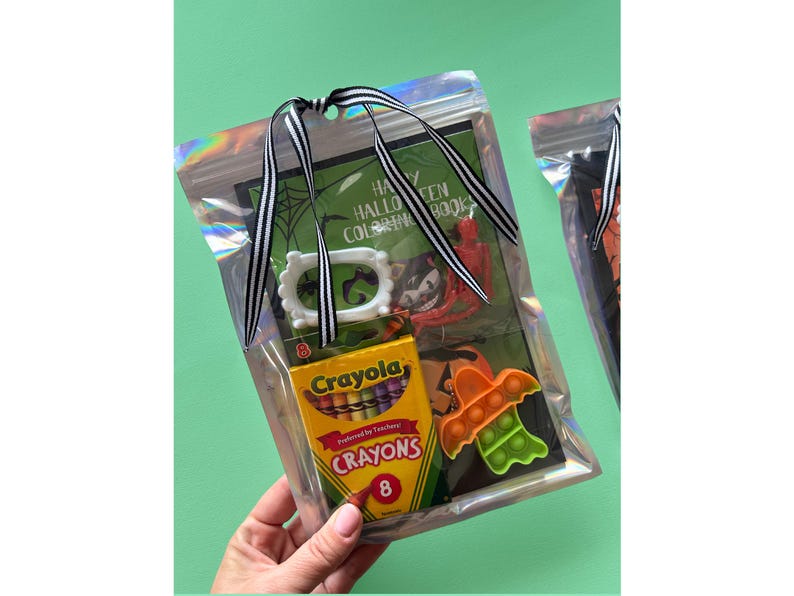 Puede incluir: Una bolsa de pl&aacute;stico iridiscente transparente atada con una cinta de rayas blancas y negras que contiene un libro para colorear de Happy Halloween, una caja peque&ntilde;a de ocho crayones Crayola, un marco de pl&aacute;stico blanco, un esqueleto de pl&aacute;stico rojo y un juguete fidget naranja con forma de murci&eacute;lago.