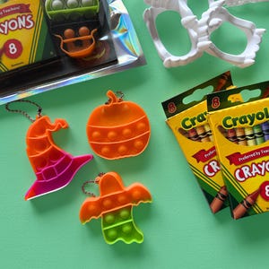 Puede incluir: Tres juguetes sensoriales de silicona con forma de sombrero de bruja, calabaza y fantasma, cada uno con un llavero de metal. Dos cajas amarillas de crayones Crayola marcadas con el n&uacute;mero 8 y colmillos de vampiro de pl&aacute;stico blanco.