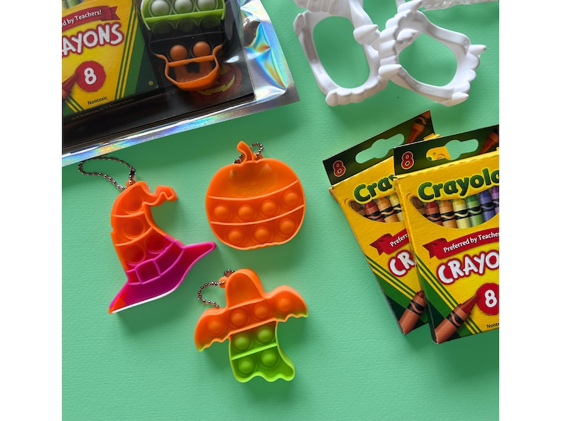 Puede incluir: Tres juguetes sensoriales de silicona con forma de sombrero de bruja, calabaza y fantasma, cada uno con un llavero de metal. Dos cajas amarillas de crayones Crayola marcadas con el n&uacute;mero 8 y colmillos de vampiro de pl&aacute;stico blanco.
