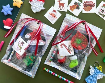 Puede incluir: Dos bolsas transparentes iridiscentes atadas con cintas rojas y blancas, cada una con un bolígrafo, un pequeño juguete de burbujas de silicona y una pegatina navideña. Las bolsas están rodeadas de objetos festivos como una estrella y un ángel.
