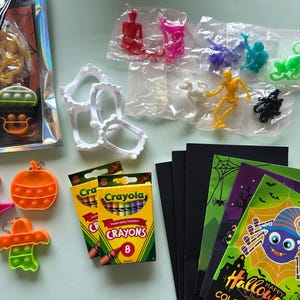 Puede incluir: Una colecci&oacute;n de art&iacute;culos para fiestas de Halloween que incluye crayones Crayola, figuras de esqueletos de pl&aacute;stico de varios colores, dientes de vampiro de pl&aacute;stico blanco, juguetes fidget de silicona naranja con formas de calabazas, fantasmas y sombreros, y libros para colorear.