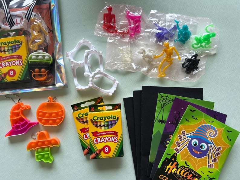 Puede incluir: Una colecci&oacute;n de art&iacute;culos para fiestas de Halloween que incluye crayones Crayola, figuras de esqueletos de pl&aacute;stico de varios colores, dientes de vampiro de pl&aacute;stico blanco, juguetes fidget de silicona naranja con formas de calabazas, fantasmas y sombreros, y libros para colorear.