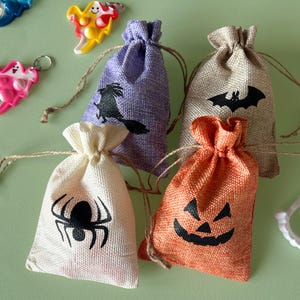 May include: Four small drawstring burlap bags in cream, purple, tan, and orange, each featuring a black Halloween-themed print of a spider, a witch, a bat, and a jack-o'-lantern face, accompanied by colorful ghost-shaped keychains and white fangs.