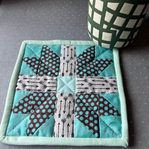 May include: A teal and brown quilted pot holder with a geometric pattern of arrows and polka dots. The pot holder has a light green border.