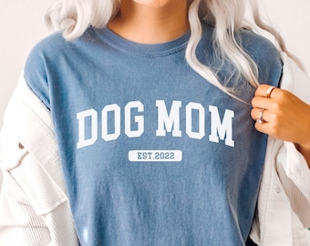 May include: A woman wearing a blue jean shirt with the text "DOG MOM EST 2022" in white. She is also wearing a white denim jacket and blue jean shorts.
