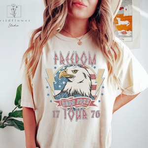 Puede incluir: Una camiseta de color crema con un gr&aacute;fico de estilo vintage de un &aacute;guila, una bandera estadounidense y el texto "FREEDOM BORN FREE 17 TOUR 76".