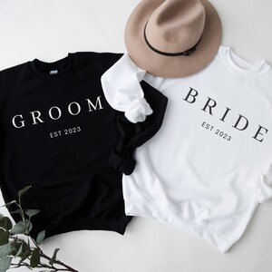 May include: Two black and white sweatshirts with the words "Groom Est 2023" and "Bride Est 2023" printed on them.